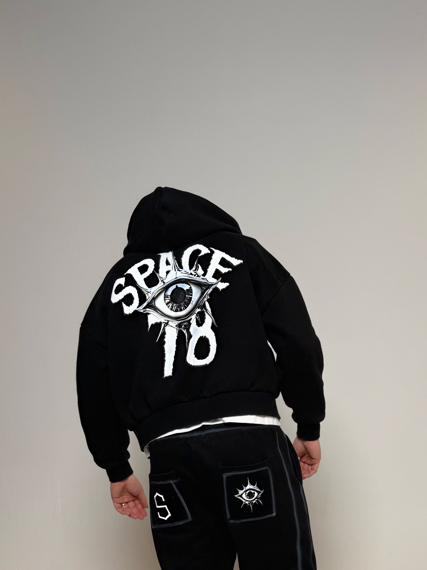 Person wearing a black hoodie and pants with 'Space 10' logo on a plain background